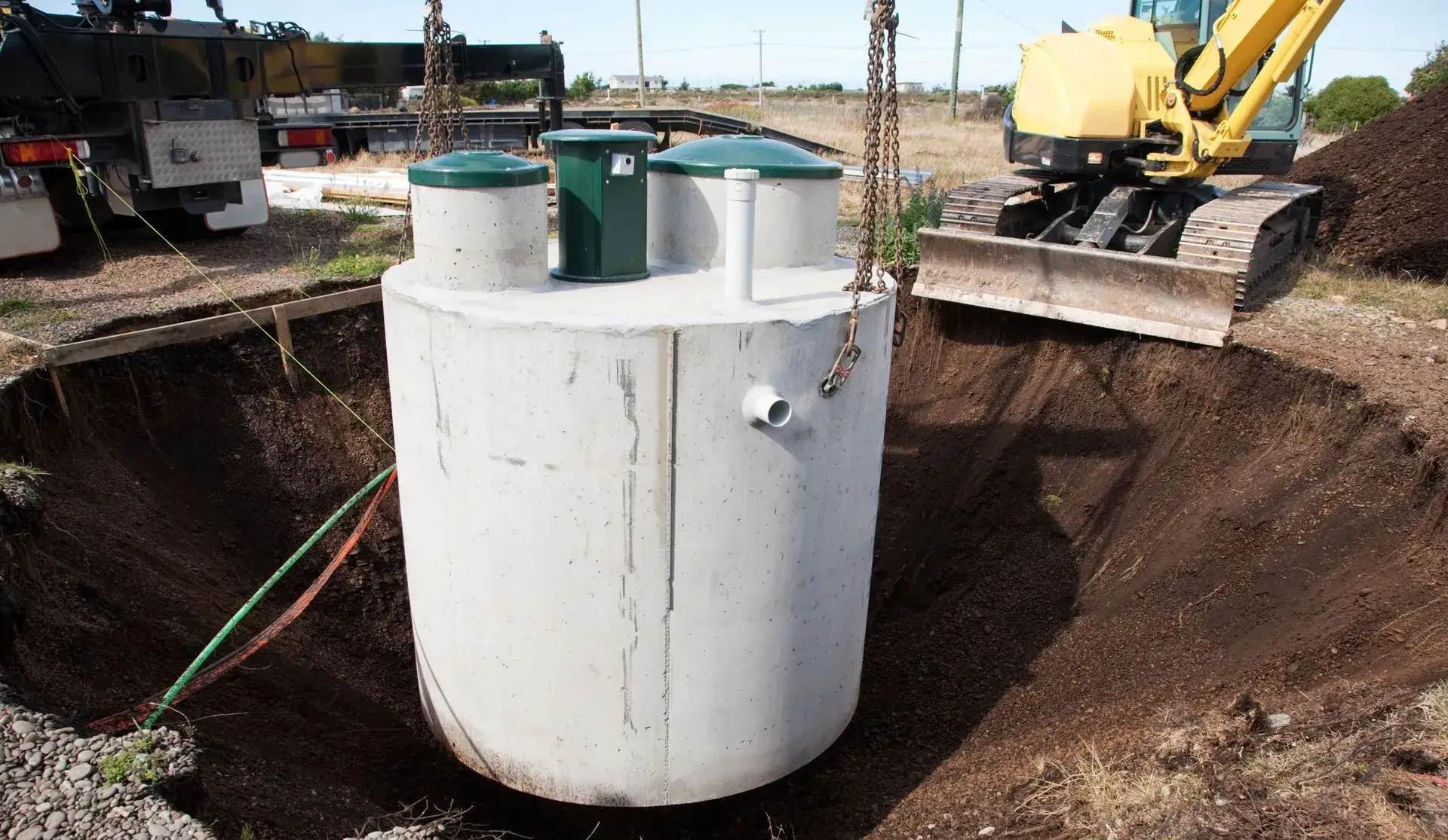 Concrete septic tank being lowered into an excavated pit by an excavator